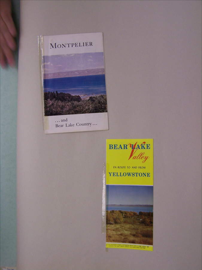 A hand is visible near an open book and a brochure on a flat surface. The book has the title "Montpelier ...and Bear Lake Country..." with an image of a landscape featuring a large body of water. The brochure is titled "Bear Lake Valley" with the subtitle "En Route To and From Yellowstone," and depicts a landscape with a body of water and foliage.