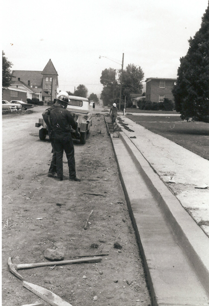 Two workers wearing helmets are standing on a dirt road next to a partially constructed curb. A vehicle is parked nearby. Construction tools are scattered on the ground. Residential houses and trees line the street.