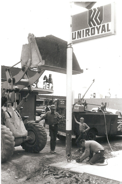 Three people are working near a signpost. One person is operating a large construction vehicle with a front loader, another person is kneeling and working on the ground near the base of the signpost, and a third person is standing nearby with one hand on a truck. The sign reads "UNIROYAL." A building in the background displays a partially visible sign with letters "HER."