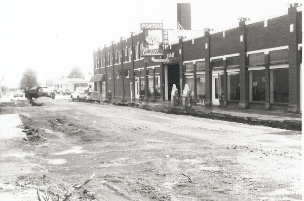 A partially constructed street in front of a row of brick buildings. The main building features signs for Pontiac, GMC, Cadillac, and Jeep, indicating it is a garage. Some construction equipment and vehicles are visible on the street, and a few workers are present.