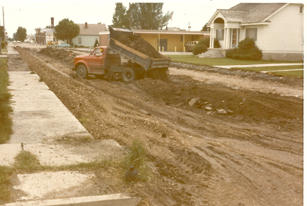 A red dump truck is unloading dirt onto a dirt road under construction in a residential area. A white house with a porch is visible on the right side. Sidewalks run along the edges of the road. In the background, a building with a sign saying "Rich" is visible, along with other houses and trees.
