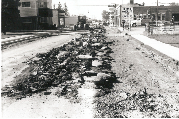 A street under construction with rubble scattered along the middle. A tractor-like vehicle is working on the road. Buildings line the street, including a visible sign for "Rich Motor Co." and "Pontiac Cadillac MC Trucks." There is a sidewalk on one side, with a single person walking.