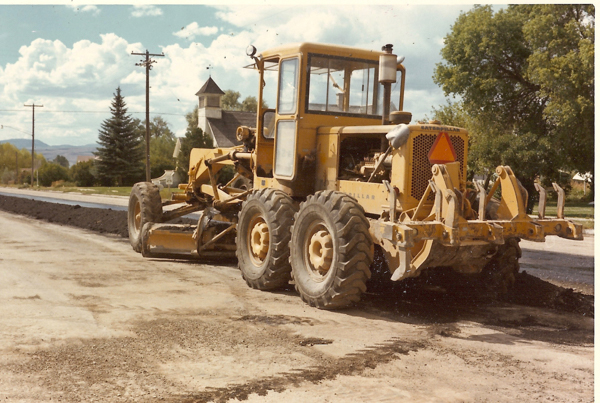 A large yellow construction vehicle, a grader, is parked on a road. The vehicle has large tires and a cab for the operator. In the background, there are trees, utility poles, and a building with a peaked roof. The sky is partially cloudy. There is a triangular orange safety sign on the back of the vehicle.