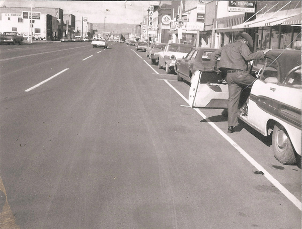 A man in a cowboy hat is stepping into a parked car along a street lined with vehicles. Businesses with signage are visible on the right, including a bank and a television store. The road stretches into the distance with a few cars driving away. There is text for a few businesses, including "EL BUFEO," "BANK," and "TELEVISION."