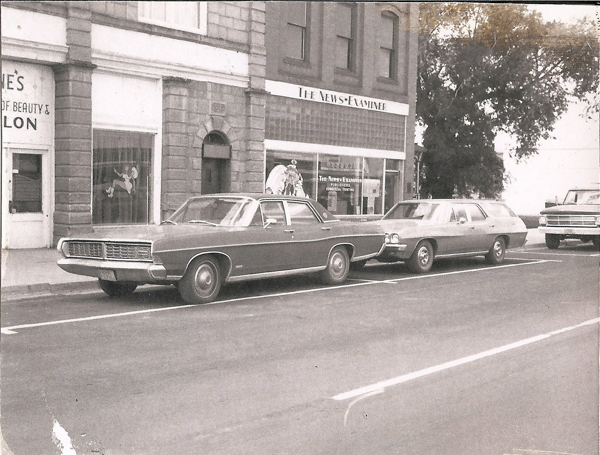 Two vintage cars are parked on a street in front of a building. One car is a sedan and the other is a station wagon. The building has signs that read "The News-Examiner" and "Louise's House of Beauty & Salon." There is a door between the signs and large windows displaying designs or decorations. Trees are visible in the background, alongside another vehicle parked further down the street.