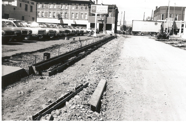 A construction site on a street with rails partially embedded in the ground. On the left, there are rows of parked cars alongside a building with the sign "L. L. Burgoyne & Sons." In the background, a large trailer truck is parked across the street. There is a sign for "AFL Used Cars" and a motorcycle parked nearby. A stop sign can be seen on the right.