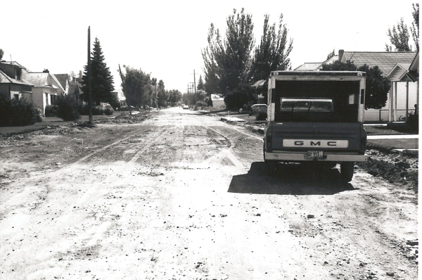 A dirt road lined with houses and trees. A GMC truck is parked on the right side of the road. The road appears uneven with visible tracks and debris.