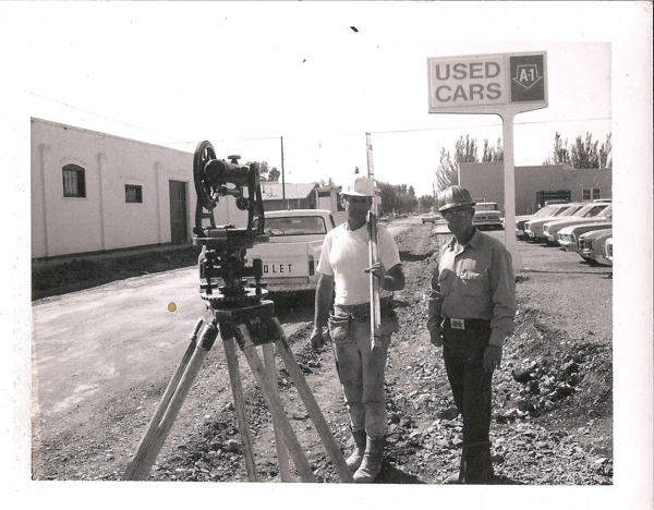 Two men wearing hard hats stand next to a tripod with survey equipment on a dirt road. One man holds a measuring rod. A pickup truck with the word "CHEVROLET" is parked nearby. A sign in the background reads "USED CARS A-1." Several cars are lined up along the side. Buildings are visible to the left and right.