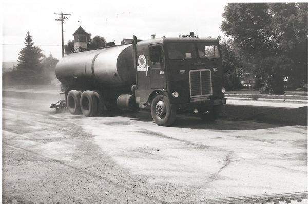 A large tanker truck labeled with the number "910" is parked on a road. There is a logo with a sun design on the side of the cab. In the background, there are trees, a telephone pole, and a building with a pointed roof. The road appears dusty.