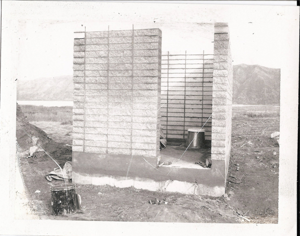 A small, unfinished building with three visible walls made of blocks and a wire support structure. The open space leads inside, where a metal container is visible on the ground. There are dirt and construction materials scattered around the site, with a pile of soil to the left and a distant mountain range in the background.