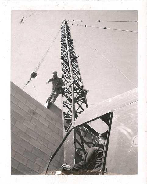 A person standing on a steel structure at a construction site, wearing work clothes and a hat. Another person is seated inside a control cabin at the base, operating machinery. The scene includes a tall metal framework and a partially constructed brick wall.