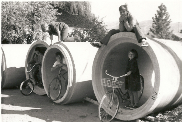 Four children are playing with bicycles around large concrete pipes. Two children are inside the pipes with bikes, one is climbing over the top, and another is sitting on top of one of the pipes, resting their chin on their hands. Trees and a building can be seen in the background.
