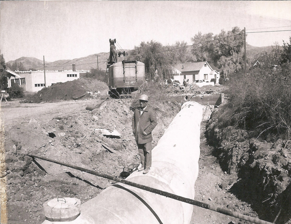 A person wearing a hard hat and coat stands next to a large pipeline in a construction area. In the background, there is construction equipment, including an excavator and a cement mixer with "TRACK PAVER" on it. There is also a pile of dirt, several buildings, and some trees. Hills can be seen in the distance.