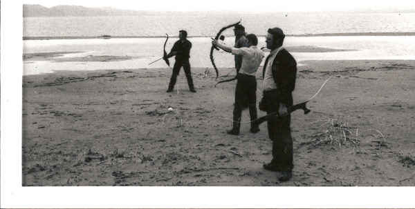 Three people standing on a sandy beach by the water, each holding a bow and aiming it. The figure in the middle draws the bowstring back, preparing to shoot. The background features distant hills or mountains across the water. Sparse vegetation and sticks are scattered on the sand.