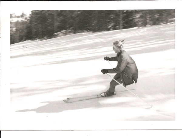A person skiing quickly down a snowy slope, wearing a jacket and gloves, with trees visible in the background.