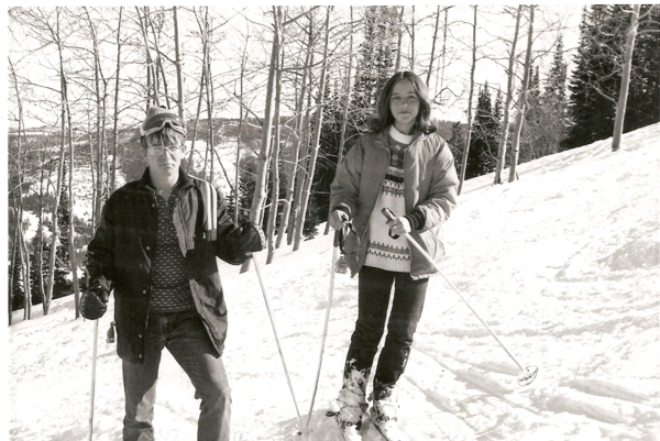 Two people standing on a snowy slope, both holding ski poles. The person on the left is wearing a jacket and goggles, while the person on the right is wearing a patterned sweater and jacket. There are bare trees and a wooded area in the background.