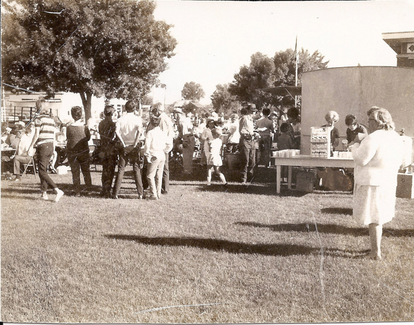 People gather outdoors on a grassy area, possibly for an event or community gathering. In the foreground, a woman holds a cup. Nearby, a table is set up with drinks and snacks, where several people are serving or helping themselves. In the background, groups of people stand and sit around, with trees and a few buildings visible.