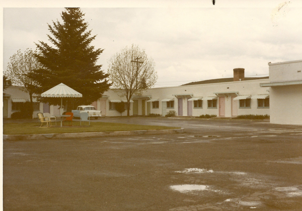 A row of single-story motel rooms, each with a different colored door and a small awning above the windows, is shown along with a parking area. In the foreground, there is an arrangement of outdoor furniture including a table and several chairs under a striped umbrella. A large tree and a car are visible near the building. The area appears to be wet, suggesting recent rain.