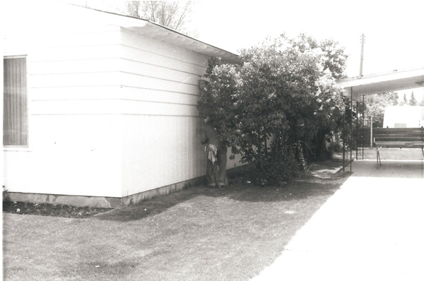 A person standing near the corner of a house partially obscured by a bush. A cloth is draped over their waist. To the right, there is a covered patio with a swing. The foreground has a grassy lawn.