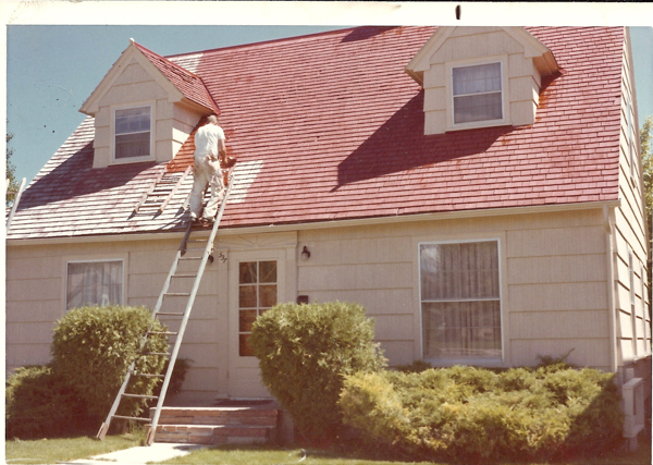A person is climbing a ladder, painting the roof of a house. The house has two dormer windows on the roof and a front door with a small window. The landscaping includes bushes near the entrance. The number "32" is visible near the door.