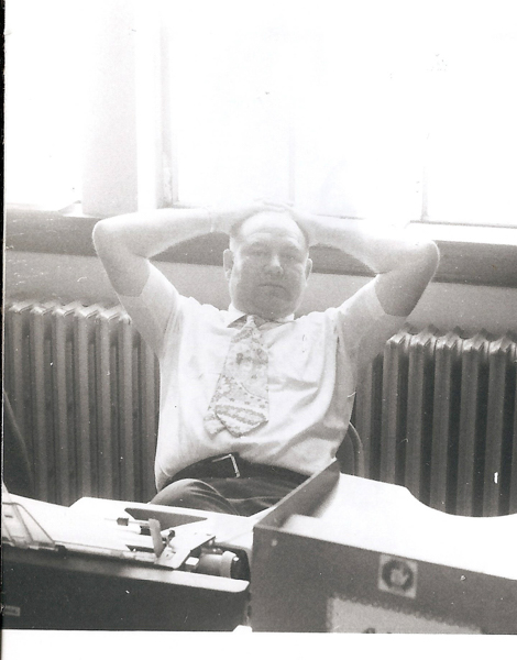 A man is sitting at a desk with his hands behind his head. There is a typewriter on the desk in front of him. He is wearing a tie and appears to be in an office setting with a radiator and window behind him.