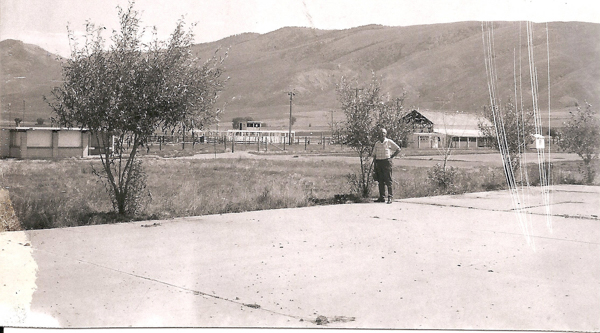 A person standing on a paved area next to a small tree. The background features open fields, a few trees, buildings, and hills in the distance. There are utility poles and fencing visible across the landscape.