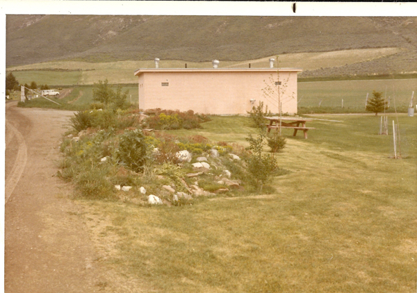 A small rectangular building with several vents on the roof is situated in a grassy area. In the foreground is a landscaped garden with various plants and rocks. To the right, a picnic table is set on the lawn. A dirt road runs alongside the garden, leading towards a distant white vehicle. In the background, rolling hills and fields stretch into the distance.