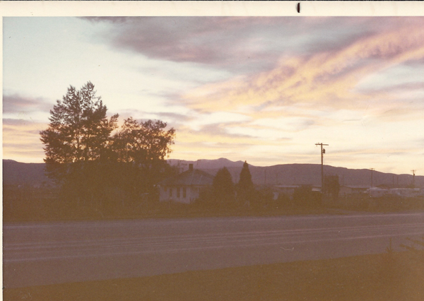A landscape with a road in the foreground. Behind the road, there are trees and a house. In the background, hills are visible beneath a sky with scattered clouds. Utility poles line the road.