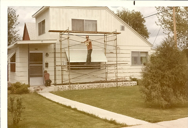 A house with two men working outside. One man is on a scaffold against the house, while the other is kneeling by the front door. The house has a sloped roof and several windows. There's a grassy lawn with a pathway leading to the front door. Visible text on the house reads "7047". Trees and shrubs are around the property.
