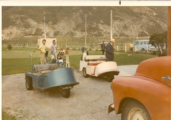 Two men are standing near a blue golf cart with golf bags on a golf course. Another man is near a pink golf cart. In the background, a man wearing a yellow shirt is putting on the green. A red vehicle is partially visible in the foreground. A blue van and mountains are in the background.