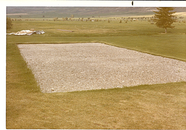 A rectangular gravel area is situated on a grassy field. In the distance, there are piles of boards and sheets of material. A single tree stands on the right, and beyond, expansive fields extend to the horizon with scattered trees and a few distant structures.