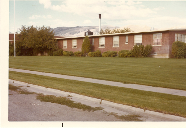 A brick building with multiple windows, with a neatly manicured lawn and sidewalk in the foreground. Bushes and trees are in front of the building, with hills visible in the background. A flagpole is on the left side.
