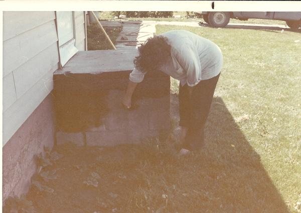 A person bending over next to a concrete structure on the side of a house, painting or applying a substance. There is grass in the background and a vehicle is partially visible.