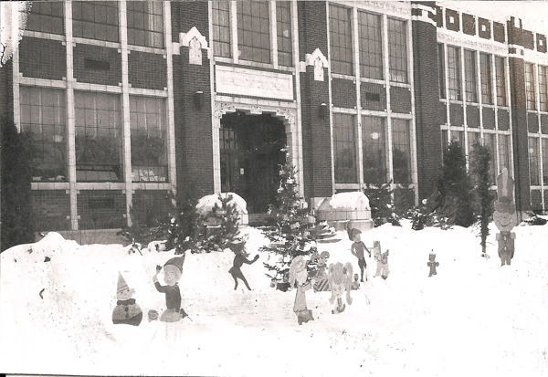 A large brick building with multiple tall windows in the background. In the foreground, there is snow on the ground and several cut-out figures of children and animals scattered throughout, suggesting a festive or playful winter scene. Small evergreen trees are positioned along the entrance steps.