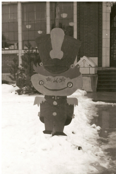 A decorative cutout of a toy soldier stands in the snow, in front of a building with large windows and a brick facade. The soldier features a large hat, a smiling face with star-shaped eyes, and a buttoned uniform. Trees are visible in the background by the building.