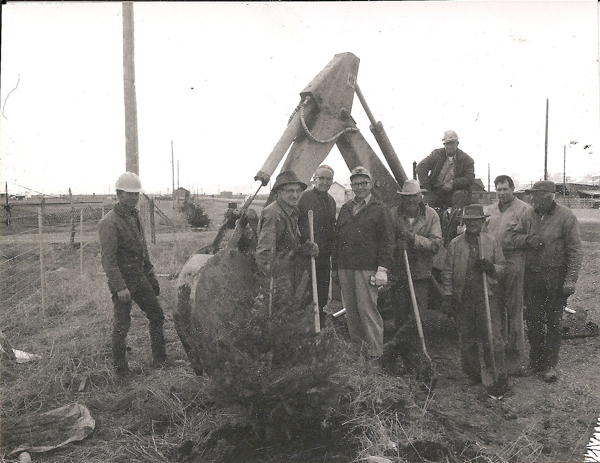 A group of men stand around a large piece of construction equipment in an open field. Some are holding shovels, and a few are wearing hard hats. A small tree is in the foreground. There are utility poles and a fence in the background.