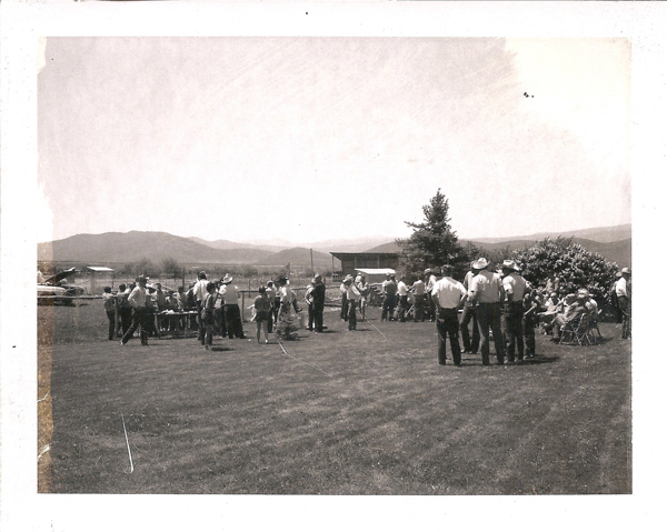 A group of people gathered on a grassy area, with mountains visible in the background. Some individuals are standing, while others are seated on chairs. A few people wear cowboy hats. There is a tree and a bush, as well as a small building and an aircraft in the distance.