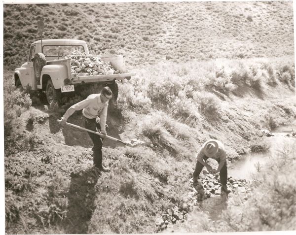 Two people working near a stream in a rural area, one using a shovel and the other picking up rocks. A truck loaded with rocks is parked nearby. The terrain is hilly with scrub vegetation. The truck's license plate reads "E 71019."