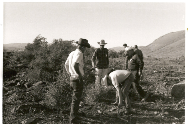 A group of people wearing hats standing outdoors on a hillside. One person is bending over, examining something on the ground. The landscape includes bushes and distant hills.