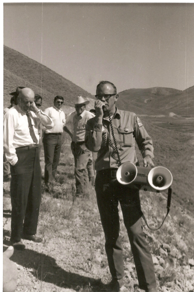 A group of men standing on a hillside. One man in the foreground is holding a megaphone and speaking into a microphone. He is wearing glasses and a uniform with patches on the sleeves. The others are dressed in shirts and ties or hats, standing slightly behind him, looking in different directions. The background consists of rolling hills.