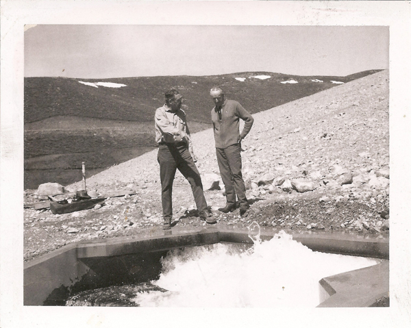 Two men stand on rocky terrain next to a concrete structure with water flowing into it. They appear to be engaged in conversation. In the background, there are hills with patches of snow. A wheelbarrow and some tools are visible on the ground.