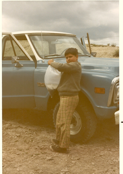 A young boy standing next to a blue truck, holding a plastic bag. He is wearing a sweater and plaid pants. The truck has the word "Custom" visible on the side. The scene is set outdoors on a cloudy day.