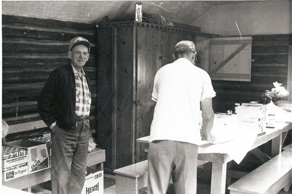A man is standing with his hands in his pockets, wearing a plaid shirt, jacket, and cap. To his left are boxes labeled "Dr Pepper," "Fanta," and "HEATH." Another man is facing away, leaning over a table covered with a tablecloth and various packaged items. The room has wooden walls and a cabinet in the background.