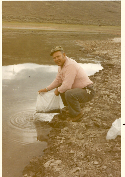 A man crouching by the edge of a body of water, holding a bag and releasing its contents into the water. The area around him is rocky and there is a small ripple in the water where the contents are being released. The background shows a hilly landscape.