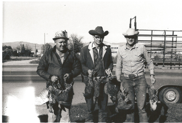 Three men stand together outdoors holding several pheasants. They are dressed in jackets and hats, with one man wearing glasses. Behind them, a sign partially visible reads “RIVER” and “MERCE.” A road and a vehicle are in the background, along with trees and hills.