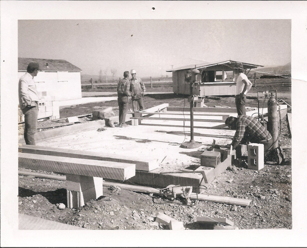 Several men are at a construction site. One man is bending down, inspecting cinder blocks and equipment. Two men are talking to each other while standing near a foundation. Another man stands off to the side, observing. In the background, there are two small buildings, and open fields are visible.