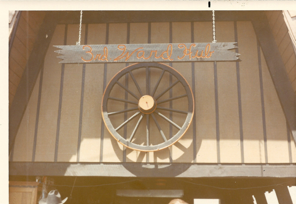 A wooden building with a large wagon wheel mounted on the wall. Above the wheel, a sign reads "3rd Ward Hub" in cursive letters. Part of a cowboy hat is visible at the bottom of the image.