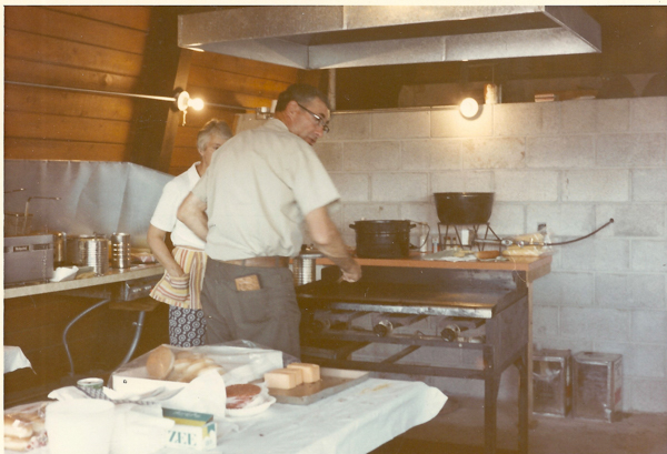 A man and a woman are in a kitchen with industrial-style equipment. The man is attending to a grill, and the woman is standing nearby with a striped apron. A table in the foreground holds bread, sliced meat, and a box labeled "ZEE." The kitchen has a concrete block wall and wooden paneling.