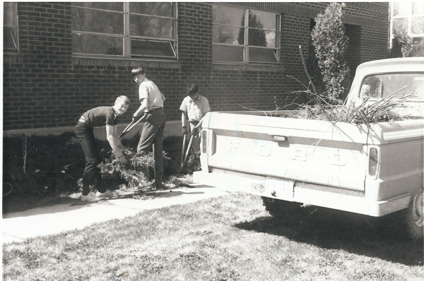 Three people are working outside in front of a brick building with large windows. They are using tools to handle branches and foliage. A pickup truck, with "FORD" written on the back, is parked nearby, carrying more branches. The scene takes place on a grassy area with a sidewalk.