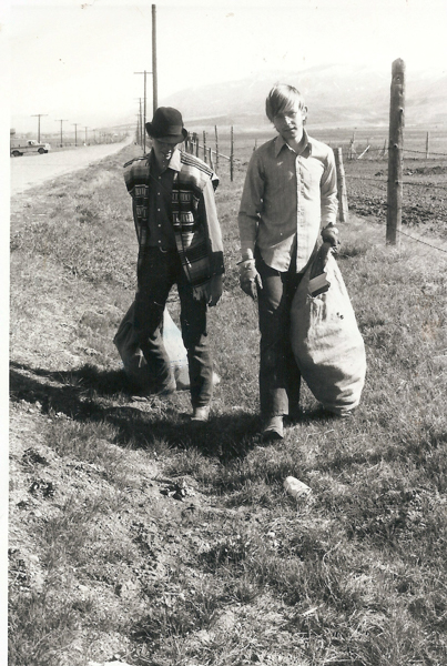 Two boys walking along a rural road, each carrying a large sack. One boy is wearing a hat and a patterned jacket, the other is wearing a button-up shirt. They are on a grassy path next to a field, with mountains in the distance. A truck is visible on the road behind them.